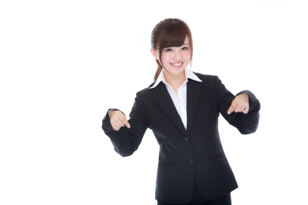 黒いスーツ姿で笑顔を浮かべ下を指差す女性の姿